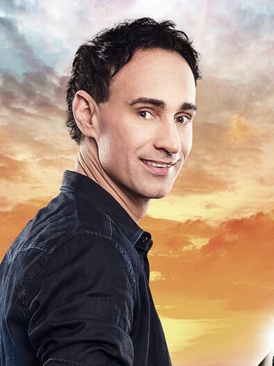 A man with a microphone stands in front of a colorful sky at sunset. He looks smilingly into the camera. | © gernot kulis