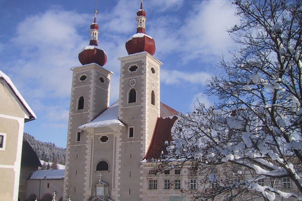 Eine Kirche mit zwei markanten Türmen und roten Kuppeln, umgeben von schneebedeckten Bäumen. Der Himmel ist klar und blau, was der frostigen Szene einen schönen Kontrast verleiht. | © Tv Murau