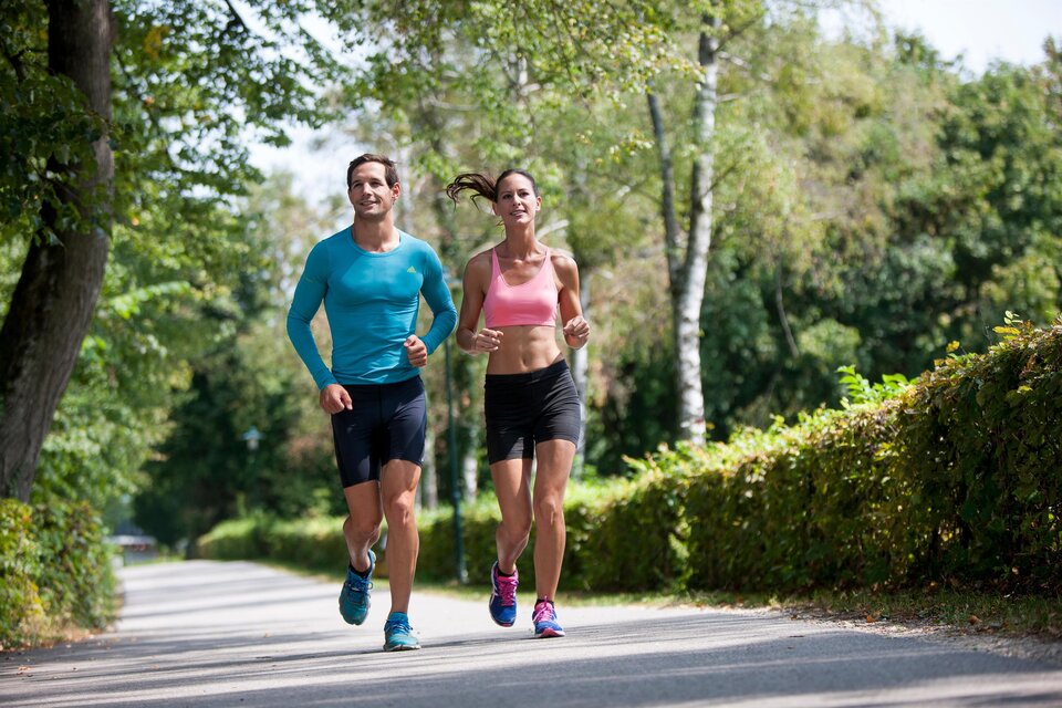 Ein aktives Paar joggt auf einem schmalen Weg umgeben von Bäumen. Es ist ein sonniger Tag und die Stimmung ist frisch und energetisch. | © (c) Region Graz - Tom Lamm