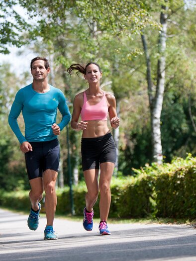 An active couple is jogging on a narrow path surrounded by trees. It is a sunny day and the mood is fresh and energetic. | © (c) Region Graz - Tom Lamm
