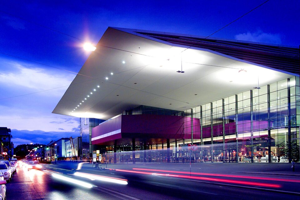 Ein modernes Gebäude bei Dämmerung, mit großflächigen Fensterfronten und einem auffälligen Dach. Verkehrslampen und Autos verleihen der Szene eine lebendige Atmosphäre.