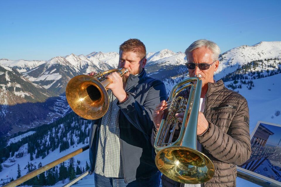 Zwei Männer spielen eine Trompete und ein Euphonium in den Bergen. Im Hintergrund sind schneebedeckte Gipfel und ein klarer Himmel zu sehen. | © Riesneralm Bergbahnen