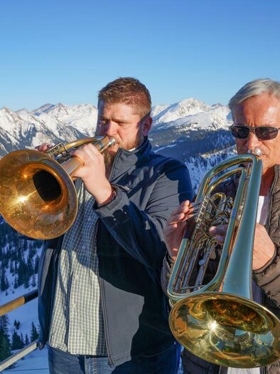 Two men are playing a trumpet and a euphonium in the mountains. In the background, snow-covered peaks and a clear sky can be seen. | © Riesneralm Bergbahnen