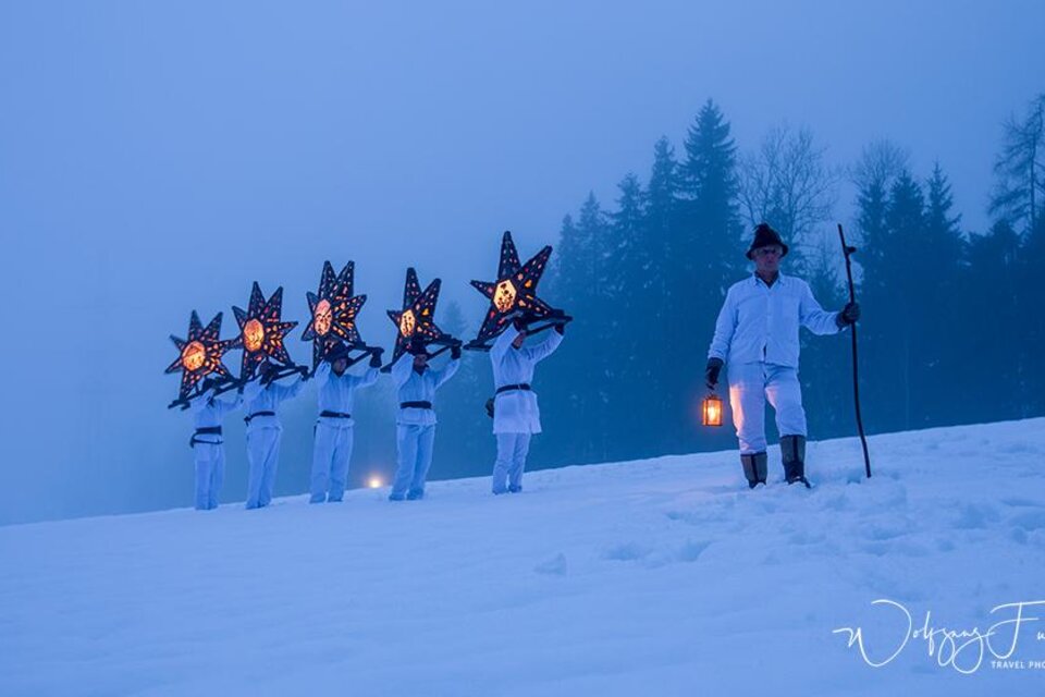 Eine Gruppe von Menschen in weißen Kleidern trägt leuchtende Sterne und marschiert durch den Schnee. Im Hintergrund sind schneebedeckte Bäume und eine neblige Atmosphäre zu sehen. | © Fuchs Wolfgang