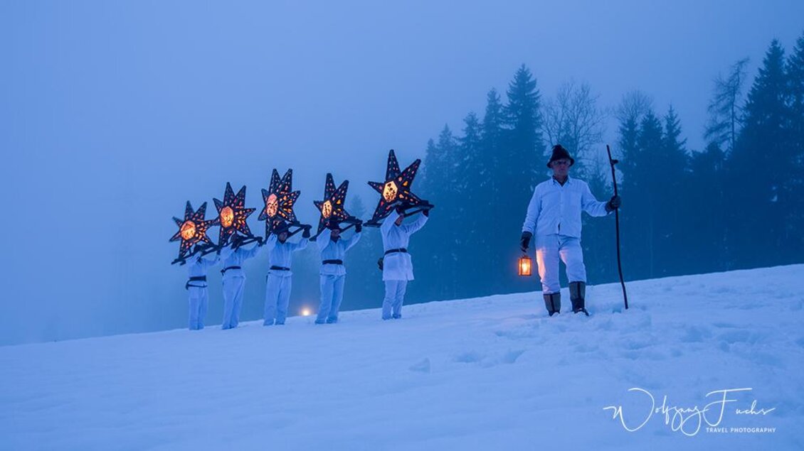 Eine Gruppe von Menschen in weißen Kleidern trägt leuchtende Sterne und marschiert durch den Schnee. Im Hintergrund sind schneebedeckte Bäume und eine neblige Atmosphäre zu sehen. | © Fuchs Wolfgang