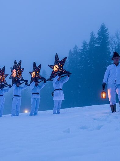 Eine Gruppe von Menschen in weißen Kleidern trägt leuchtende Sterne und marschiert durch den Schnee. Im Hintergrund sind schneebedeckte Bäume und eine neblige Atmosphäre zu sehen. | © Fuchs Wolfgang