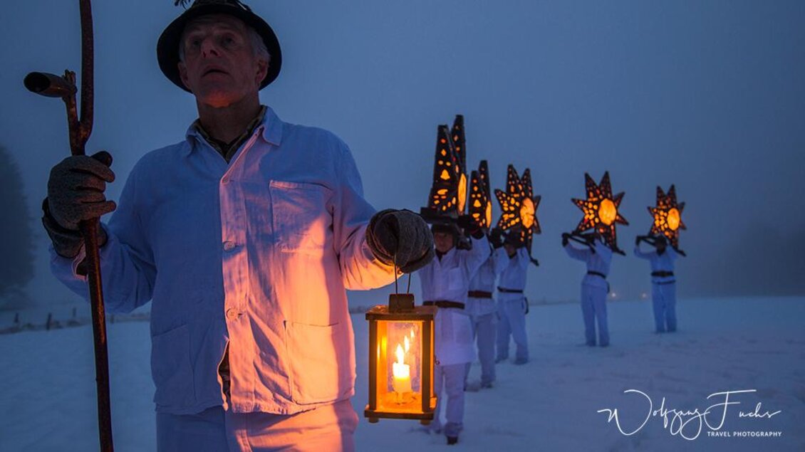 Eine Gruppe von Menschen in weißen Kleidung trägt Laternen in einer verschneiten Landschaft. Im Vordergrund hält ein Mann mit einem Stock eine helle Laterne. | © Wolfgang Fuchs