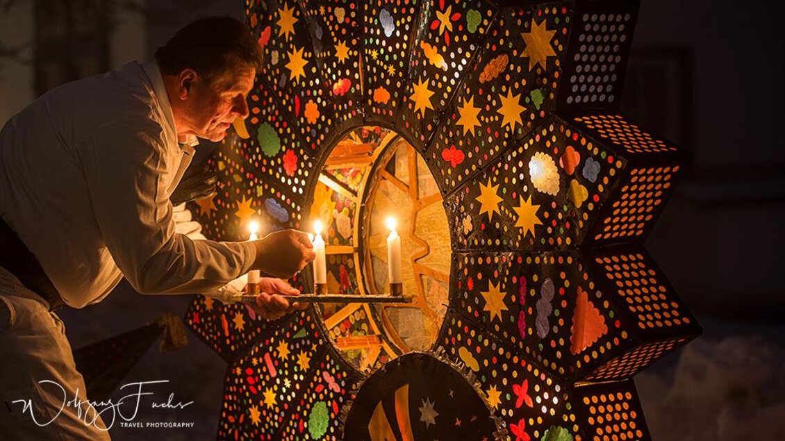 Ein Mann zündet Kerzen an einem farbenfrohen, dekorativen Objekt an. Die Szene ist in sanftem Licht gehalten und vermittelt eine festliche Atmosphäre. | © Wolfgang Fuchs