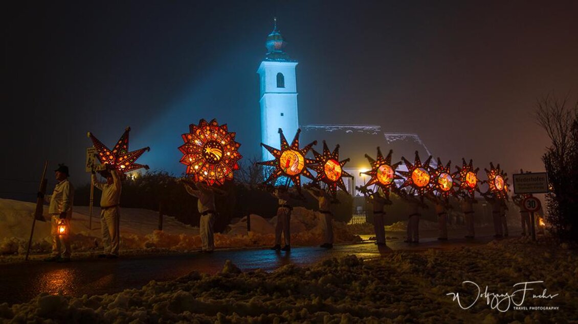 Eine Gruppe von Menschen trägt leuchtende Sternlaternen vor einer beleuchteten Kirche in einer nebligen Umgebung. Der sanfte Schnee am Boden verstärkt die festliche Stimmung. | © Wolfgang Fuchs