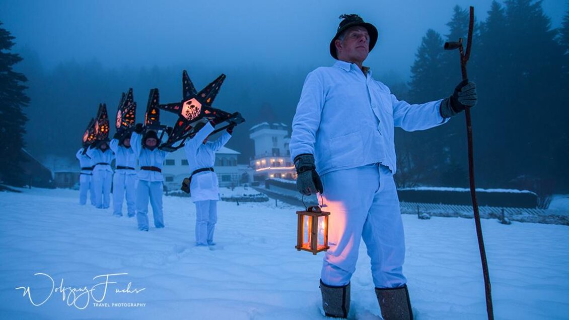 Eine Gruppe von Menschen in weißen Anzügen geht durch den Schnee, während sie leuchtende Sterne tragen. Ein Anführer hält eine Laterne und schaut ernst in die Ferne. | © Wolfgang Fuchs