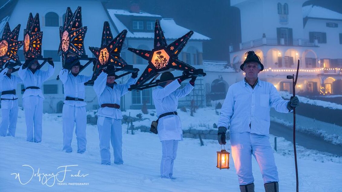 Eine Gruppe von Menschen in weißen Anzügen trägt große, leuchtende Sterne und geht durch den Schnee. Im Hintergrund sind verschneite Gebäude und eine winterliche Atmosphäre zu sehen. | © Wolfgang Fuchs