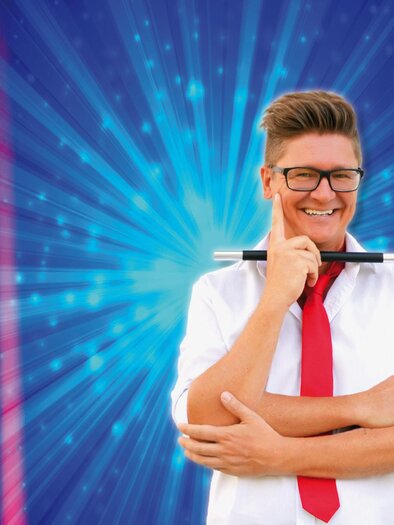 A smiling young man is wearing a red tie and holding a magic wand. The background features colorful, radiant light effects. | © Zauberkünstler Gabriel
