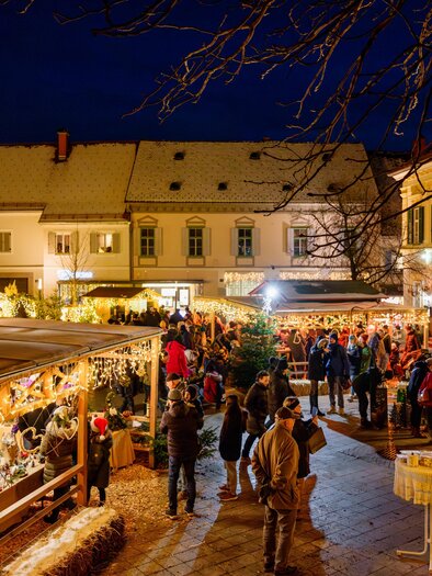 Christmas market from the side with visitors at the mulled wine stand and atmospheric lighting in the evening. | © Tourismusverband Oststeiermark