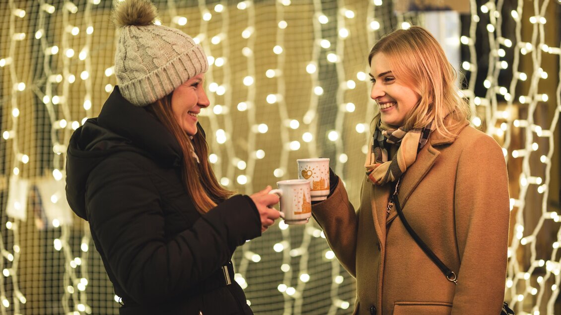 Zwei Frauen stehen lächelnd vor einer beleuchteten Hintergrundkulisse. Sie stoßen mit ihren Tassen an und genießen die festliche Stimmung. | © Tourismusverband Oststeiermark