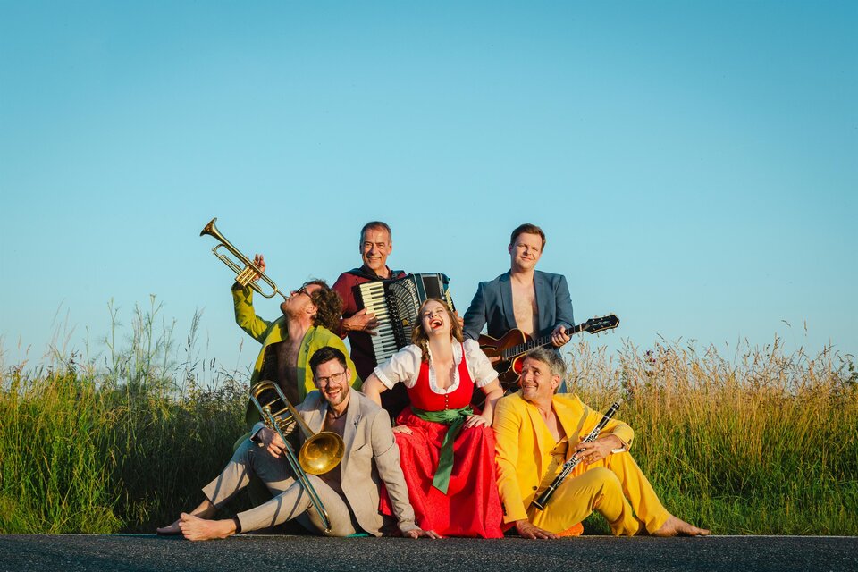 Sechs Musikerinnen und Musiker posieren fröhlich auf einer Landstraße vor hohem Gras und blauem Himmel: eine Frau in rotem Dirndl sitzt in der Mitte, umgeben von Männern in farbenfrohen Anzügen mit Trompete, Posaune, Klarinette, Akkordeon und Gitarre; die Gruppe lacht und wirkt ausgelassen. | © Astrid Bartl