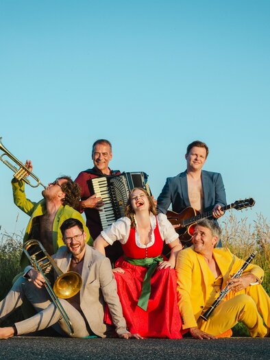 Sechs Musikerinnen und Musiker posieren fröhlich auf einer Landstraße vor hohem Gras und blauem Himmel: eine Frau in rotem Dirndl sitzt in der Mitte, umgeben von Männern in farbenfrohen Anzügen mit Trompete, Posaune, Klarinette, Akkordeon und Gitarre; die Gruppe lacht und wirkt ausgelassen. | © Astrid Bartl