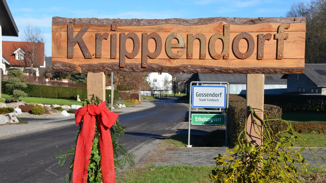 Ein Holzschild mit dem Schriftzug "Krippendorf" begrüßt die Besucher. Im Hintergrund sind Wohnhäuser und eine ruhige Straße zu sehen. | © Gossendorfer Krppenweg