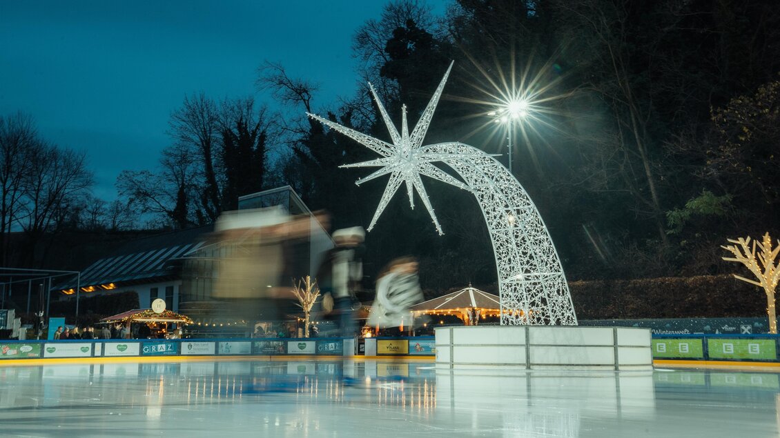 Eine Eisbahn bei Nacht mit einer leuchtenden Sternen-Skulptur. Im Hintergrund sind festlich beleuchtete Bäume und Hütten sichtbar. | © Region Graz-studio draussen