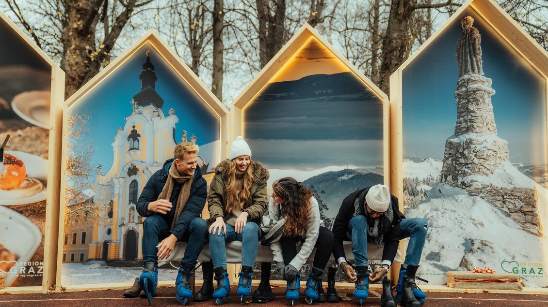 Eine Gruppe von Freunden zieht sich Schlittschuhe an, während sie vor großen Bildern von Graz sitzen. Die Atmosphäre ist gemütlich und einladend. | © Region Graz-studio draussen