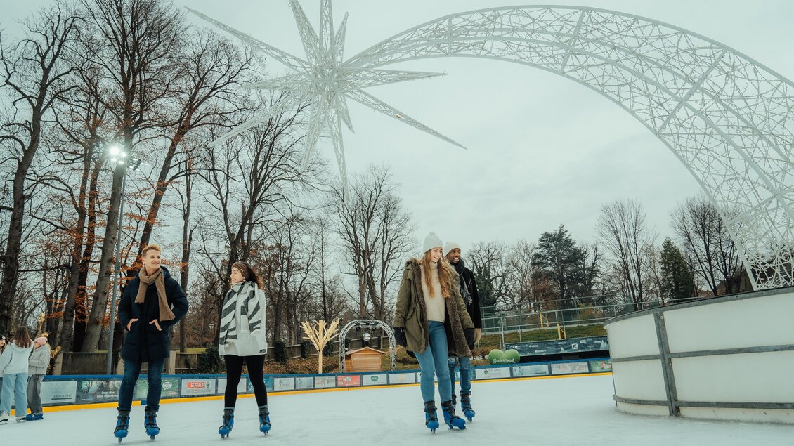 Ein Eislaufplatz mit mehreren Personen, die auf dem Eis skaten. Im Hintergrund sind Bäume und eine dekorative Struktur zu sehen. | © Region Graz-studio draussen