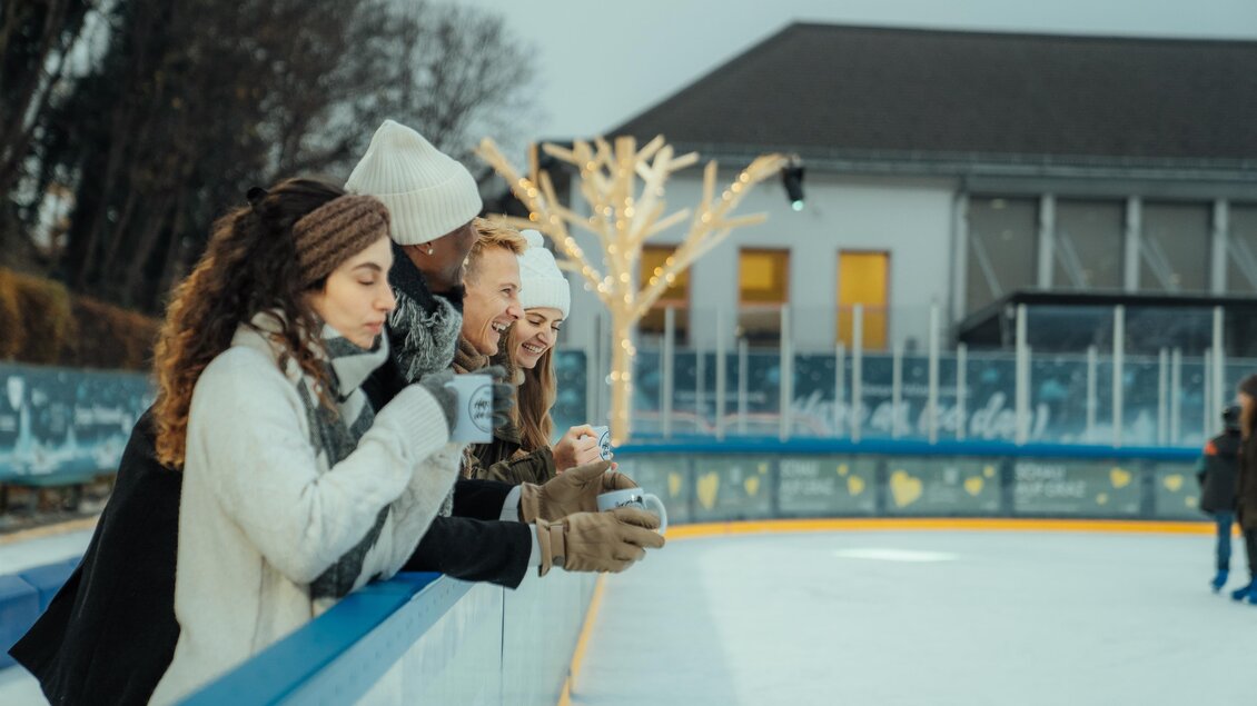 Eine Gruppe von Freunden steht an der Kante einer Eisbahn und genießt warme Getränke. Im Hintergrund sind festlich dekorierte Bäume und ein Gebäude zu sehen. | © Region Graz-studio draussen