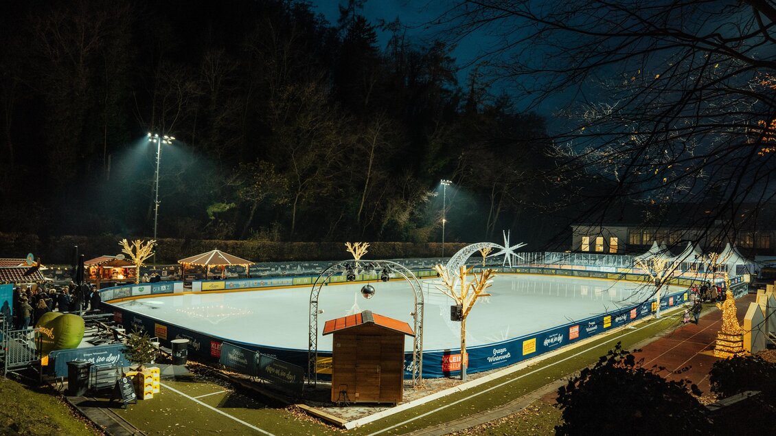 Eine beleuchtete Eisbahn bei Nacht, umgeben von Bäumen. Es gibt Stände für Snacks und Getränke in der Nähe. | © Region Graz-studio draussen