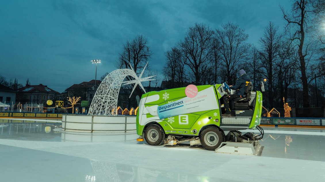 Ein Eisbahn mit einer Eismaschine, die das Eis glättet. Im Hintergrund sind beleuchtete Dekorationen und Bäume zu sehen. | © Region Graz-studio draussen
