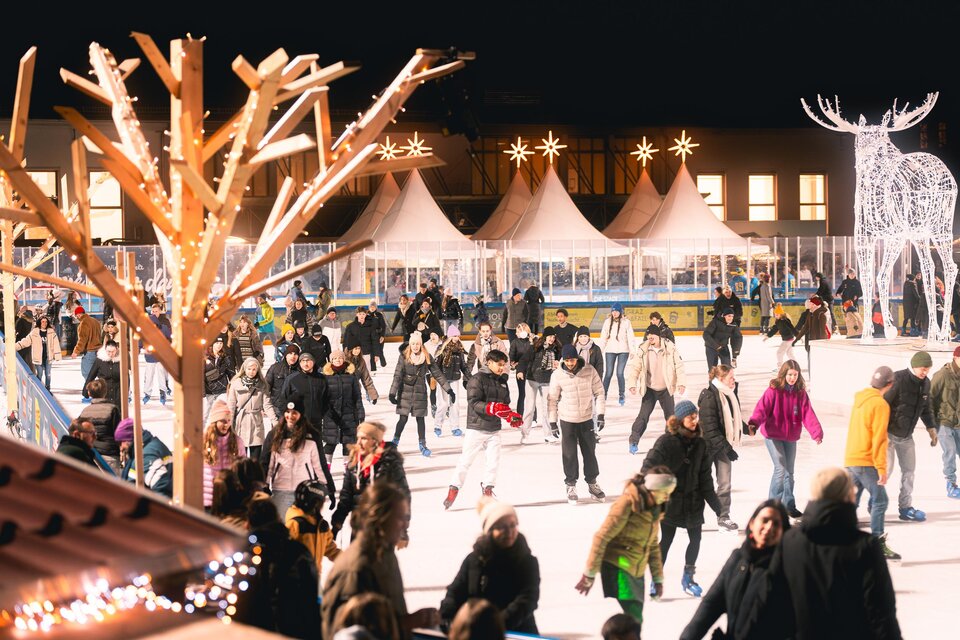 Die belebte Eisfläche der Winterwelt in der Abenddämmrung mit vielen Schlittschuhläufern. Festliche Lichter und die Gastronomie mit Genussmarkt in den Zelten im Hintergrund. | © Simon Kuro
