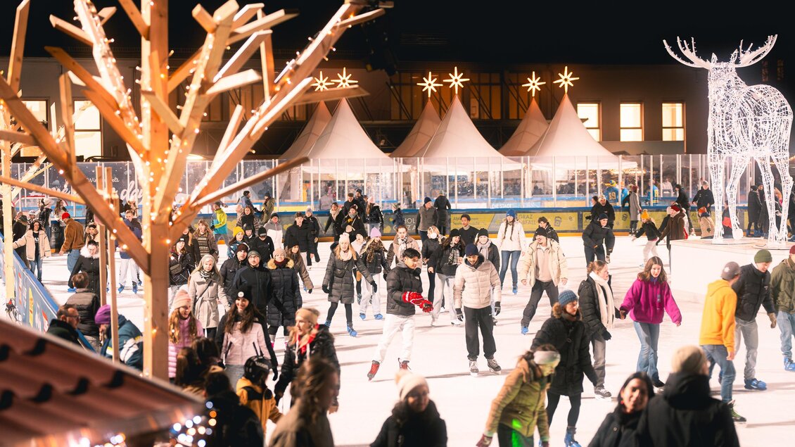 Die belebte Eisfläche der Winterwelt in der Abenddämmrung mit vielen Schlittschuhläufern. Festliche Lichter und die Gastronomie mit Genussmarkt in den Zelten im Hintergrund. | © Simon Kuro