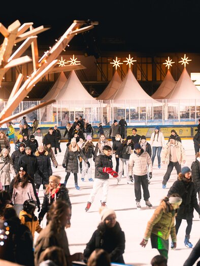 The bustling ice rink of the winter world at twilight with many ice skaters. Festive lights and the gastronomy with a gourmet market in the tents in the background. | © Simon Kuro