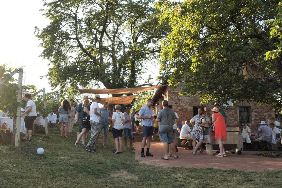 Eine lebhafte Feier im Freien mit vielen Menschen um ein rustikales Gebäude. Die Szene wird von Sonnenlicht und grünem Gras umrahmt. | © Rupert Bruchmann