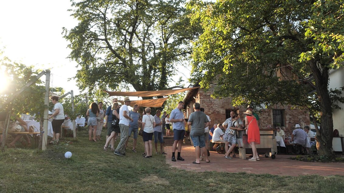 Eine lebhafte Feier im Freien mit vielen Menschen um ein rustikales Gebäude. Die Szene wird von Sonnenlicht und grünem Gras umrahmt. | © Rupert Bruchmann