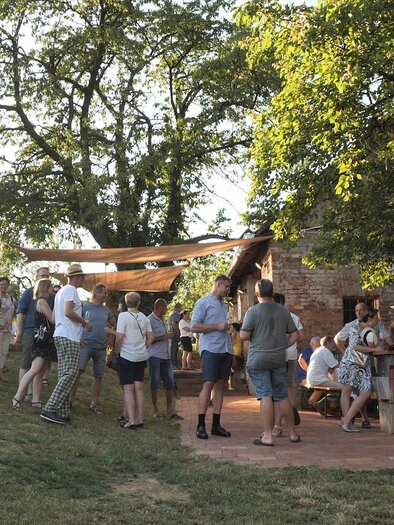 A lively outdoor celebration with many people around a rustic building. The scene is framed by sunlight and green grass. | © Rupert Bruchmann
