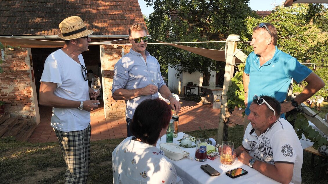Eine gesellige Runde von Männern, die an einem Tisch im Freien stehen und sich unterhalten. Im Hintergrund sind Bäume und ein Gebäude zu sehen. | © Rupert Bruchmann