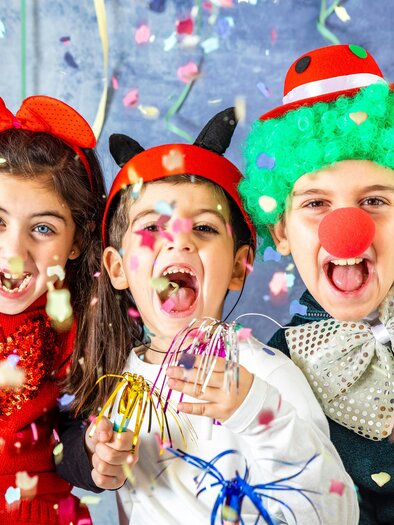 Three cheerful children are celebrating Carnival with colorful hats and confetti. They are laughing and holding colorful party items in their hands. | © TV Südsteiermark - Victor