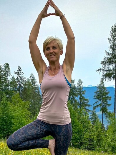 A woman is performing a yoga pose in a green meadow with trees in the background. The surroundings are calm and connected to nature. | © biYou Ecoquartier