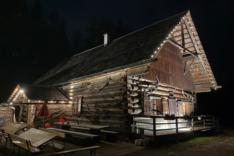 Ein rustikales Holzhaus, das nachts beleuchtet ist. Die Umgebung ist dunkel, und es gibt Sitzgelegenheiten im Freien. | © Eselalm