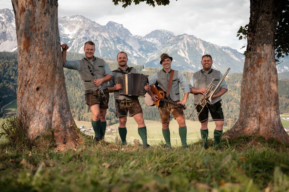 Vier Musiker in traditioneller bayerischer Tracht stehen vor einer malerischen Berglandschaft. Sie halten verschiedene Instrumente und lächeln in die Kamera. | © „Roßfeld Musi"