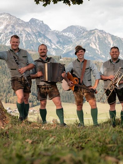 Vier Musiker in traditioneller bayerischer Tracht stehen vor einer malerischen Berglandschaft. Sie halten verschiedene Instrumente und lächeln in die Kamera. | © „Roßfeld Musi"