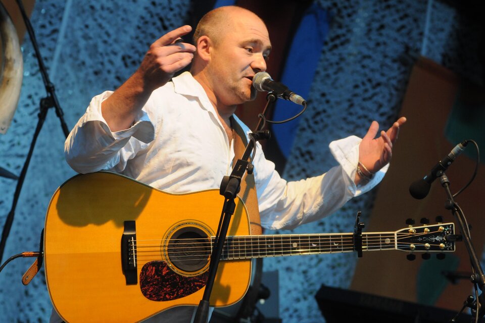 Ein Musiker spielt Gitarre und spricht ins Mikrofon. Er trägt ein weißes Hemd und steht auf einer Bühne. | © Harry Ahamer 