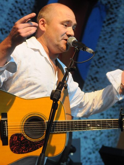 Ein Musiker spielt Gitarre und spricht ins Mikrofon. Er trägt ein weißes Hemd und steht auf einer Bühne. | © Harry Ahamer 