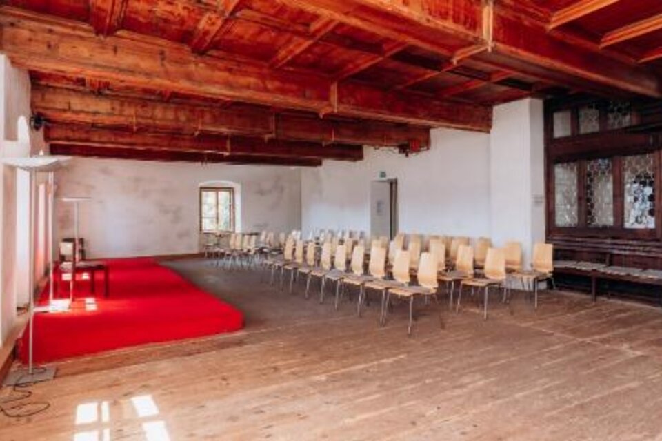Heller historischer Raum mit Holzboden, Holzbalkendecke, kleiner roter Bühne und in Reihen aufgestellten hellen Stühlen. | © Pfarrhof Pürgg