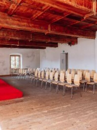 Bright historical room with wooden floors, wooden beam ceilings, a small red stage, and light-colored chairs arranged in rows. | © Pfarrhof Pürgg