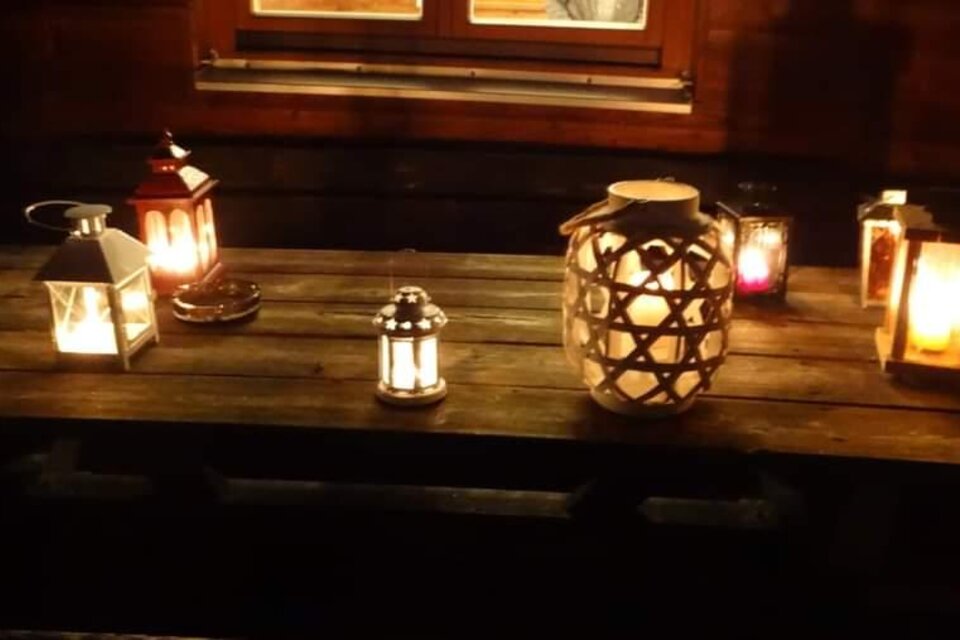 Eine rustikale Holzoberfläche mit mehreren Laternen, die in der Dunkelheit leuchten. Im Hintergrund ist ein Fenster sichtbar, das warmes Licht ausstrahlt. | © ©Thermen-&Vulkanland/Christian Rossmann