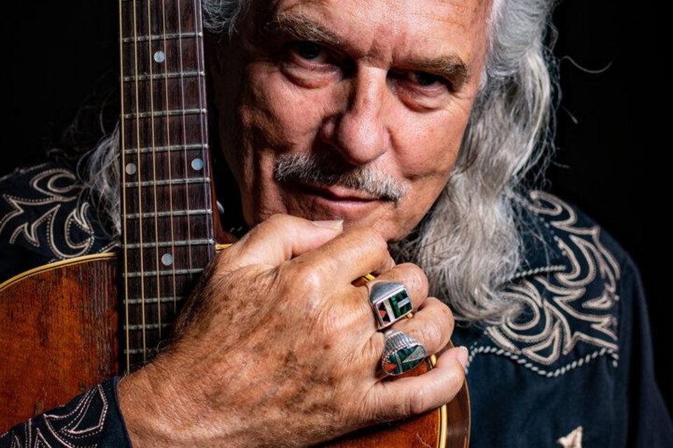 Ein älterer Mann mit langen, grauen Haaren hält eine Gitarre. Er trägt einen schwarzen Hemd mit einem auffälligen Muster und schaut direkt in die Kamera. | © Hans Theessink