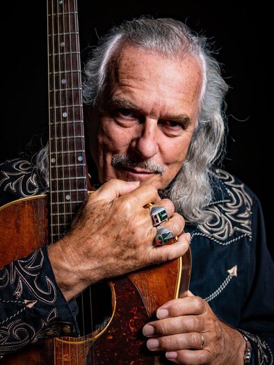 An older man with long, gray hair is holding a guitar. He is wearing a black shirt with a striking pattern and is looking directly at the camera. | © Hans Theessink