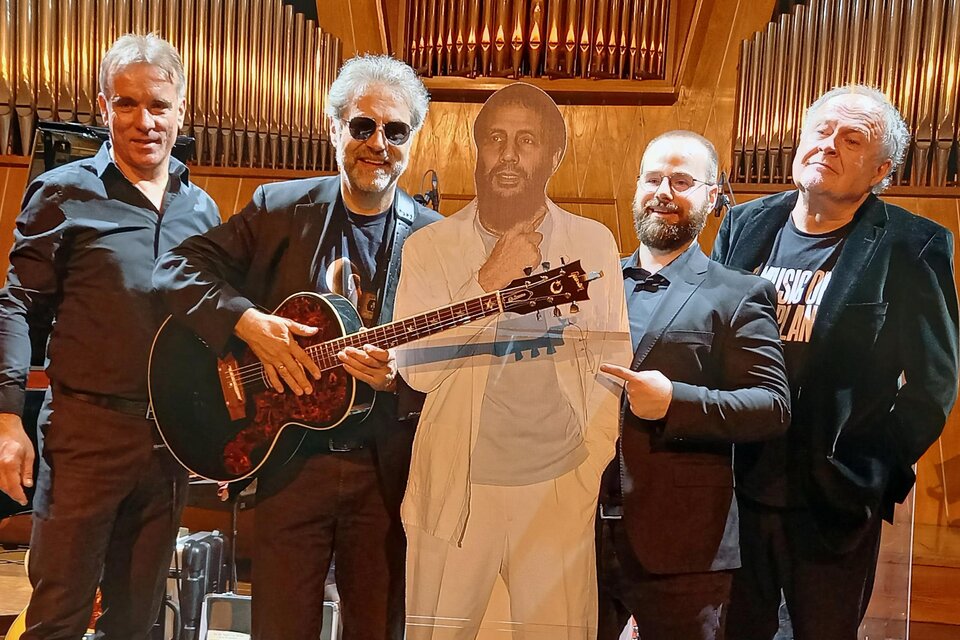 Fünf Musiker stehen zusammen auf einer Bühne, einer davon hält eine Gitarre. Neben ihnen steht eine Pappfigur eines Mannes in einem weißen Anzug. | © Harald Aigner