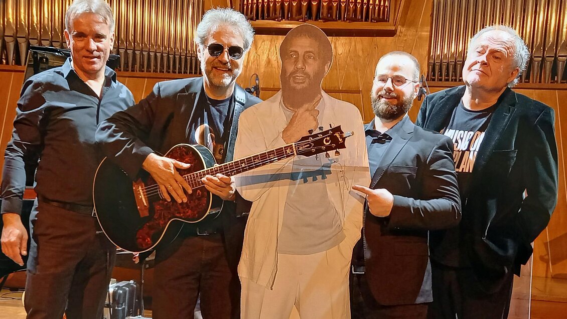 Fünf Musiker stehen zusammen auf einer Bühne, einer davon hält eine Gitarre. Neben ihnen steht eine Pappfigur eines Mannes in einem weißen Anzug. | © Harald Aigner