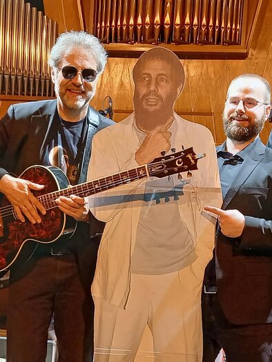 Fünf Musiker stehen zusammen auf einer Bühne, einer davon hält eine Gitarre. Neben ihnen steht eine Pappfigur eines Mannes in einem weißen Anzug. | © Harald Aigner