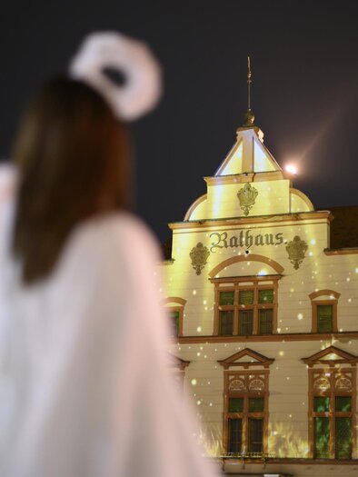 A person in a white robe stands in front of a beautifully decorated building with festive lights. The sky is dark and the atmosphere is Christmas-like. | © Tourismus Oststeiermark